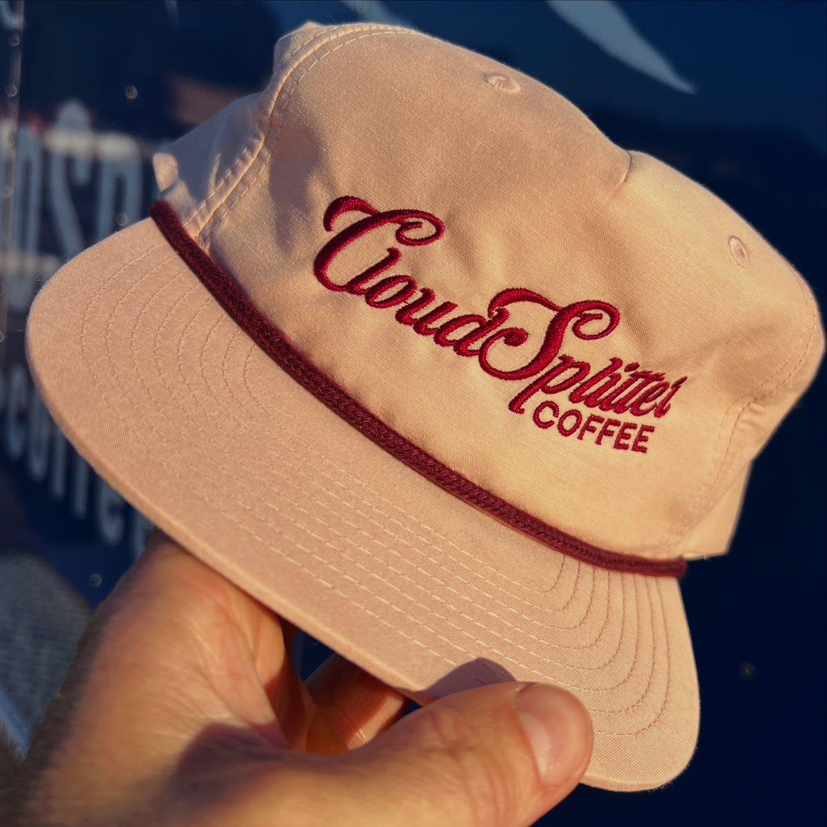 Beige 5-panel cap with maroon 'CloudSplitter Coffee' text held by a hand against a blurred background