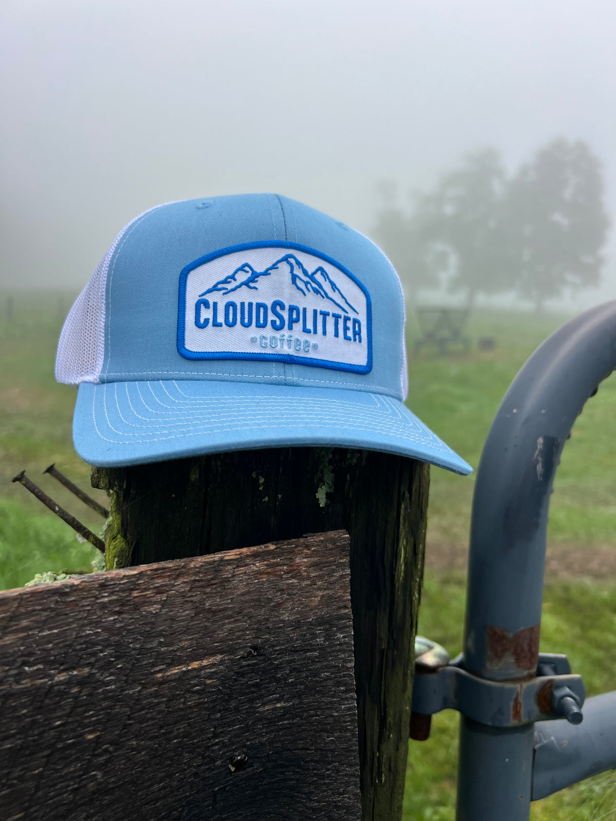 A light blue trucker hat with a white mesh back and a front patch reading CLOUDSPLITTER COFFEE, resting on a wooden fence post in a misty outdoor field.