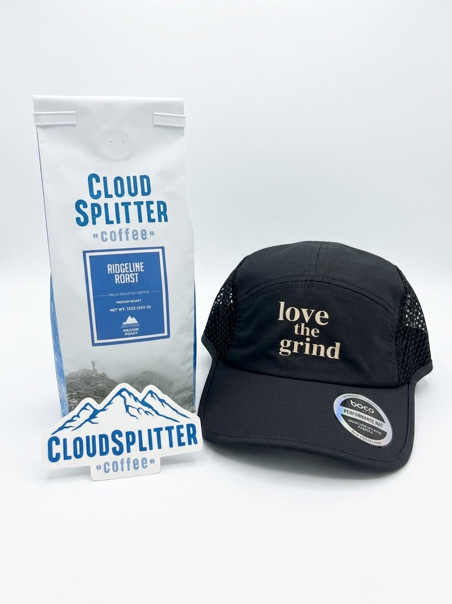A black runners cap with 'love the grind' embroidered in tan on the front, displayed next to a bag of Cloud Splitter Coffee and a small Cloud Splitter logo sticker  on a white background.