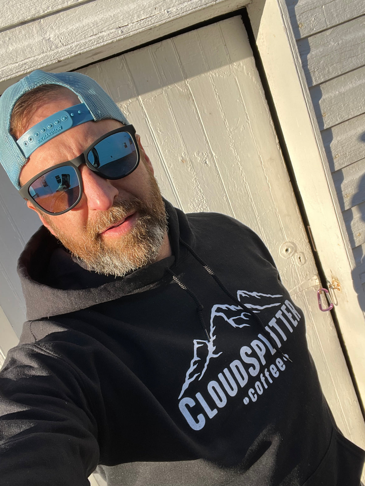 Person wearing a black hoodie with CloudSplitter Coffee text, wearing sunglasses, standing in front of a building.